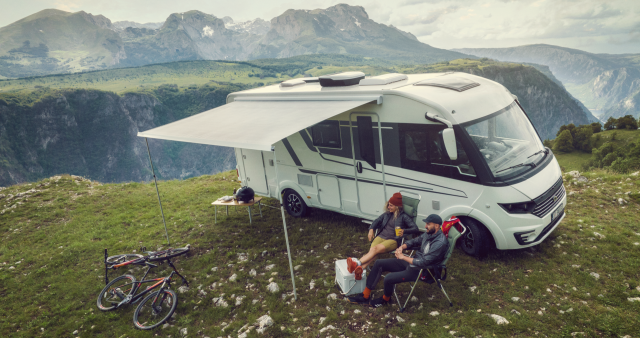 Dette bildet viser en moderne, hvit bobil parkert på en fjelltopp med en fantastisk utsikt over et frodig dalføre og majestetiske fjell.