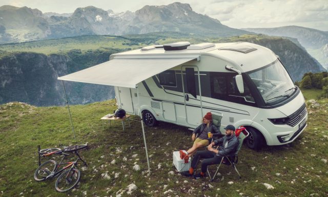 Dette bildet viser en moderne, hvit bobil parkert på en fjelltopp med en fantastisk utsikt over et frodig dalføre og majestetiske fjell.